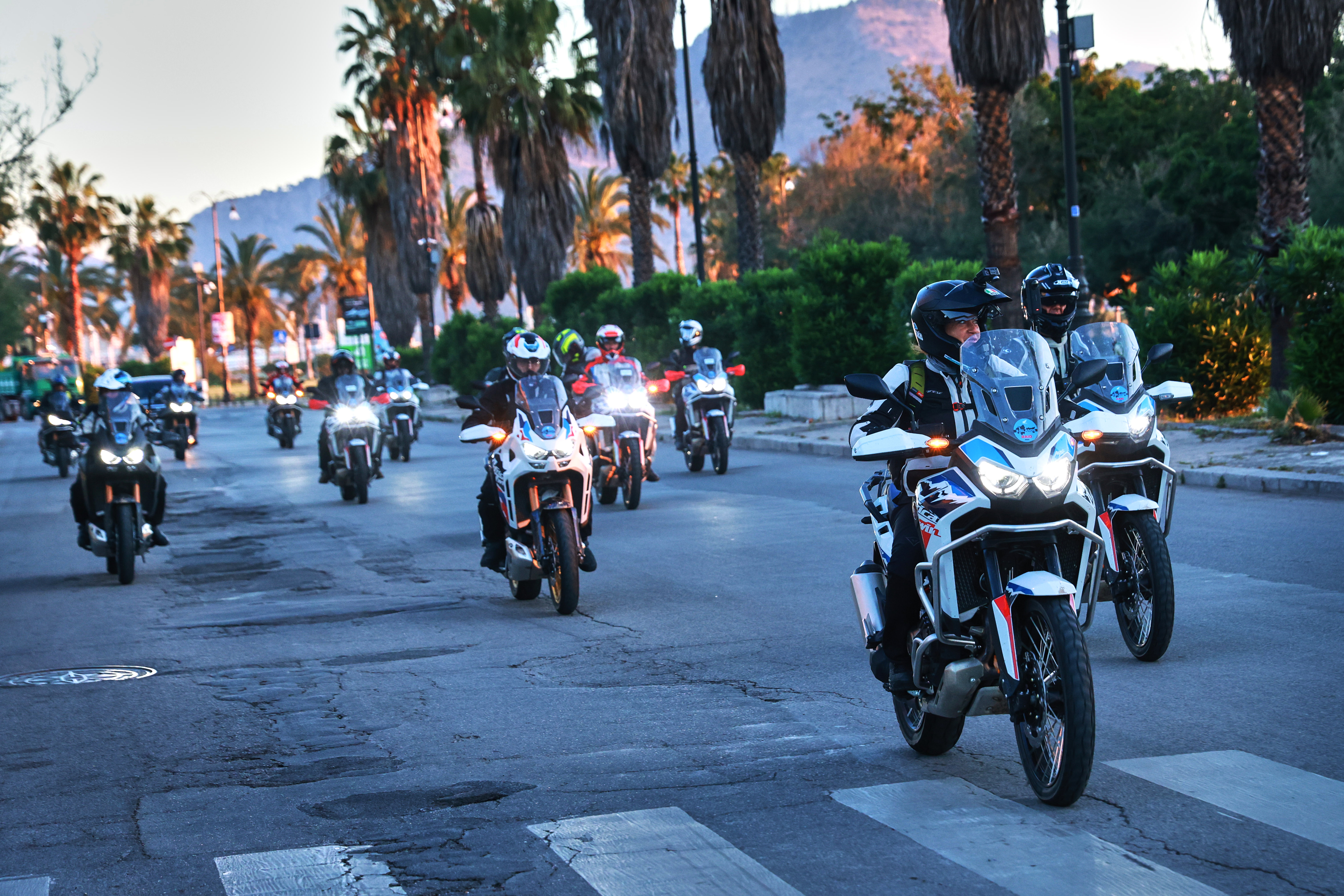 Una colonna di moto Honda Africa Twin che percorre una strada fiancheggiata da palme durante il tramonto.